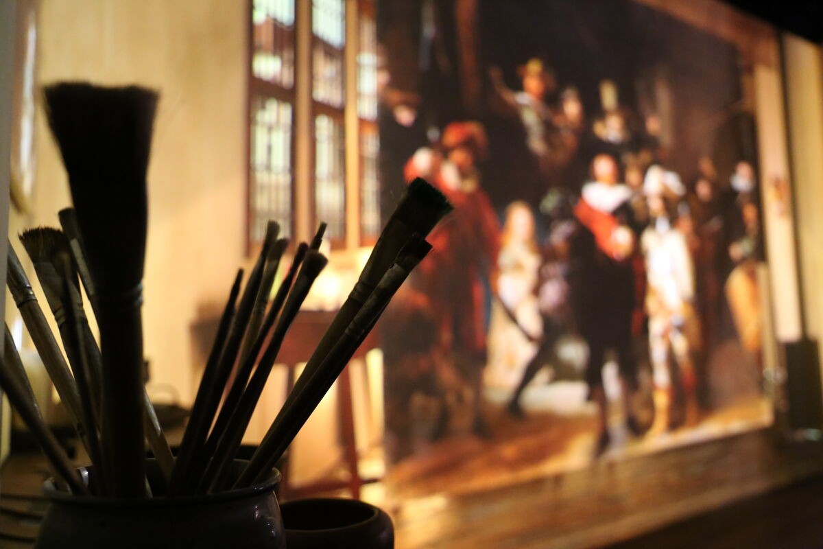 Een close-up van een bundel verfkwasten. In de achtergrond, niet in focus, is een stuk van De Nachtwacht van Rembrandt van Rijn te zien. 