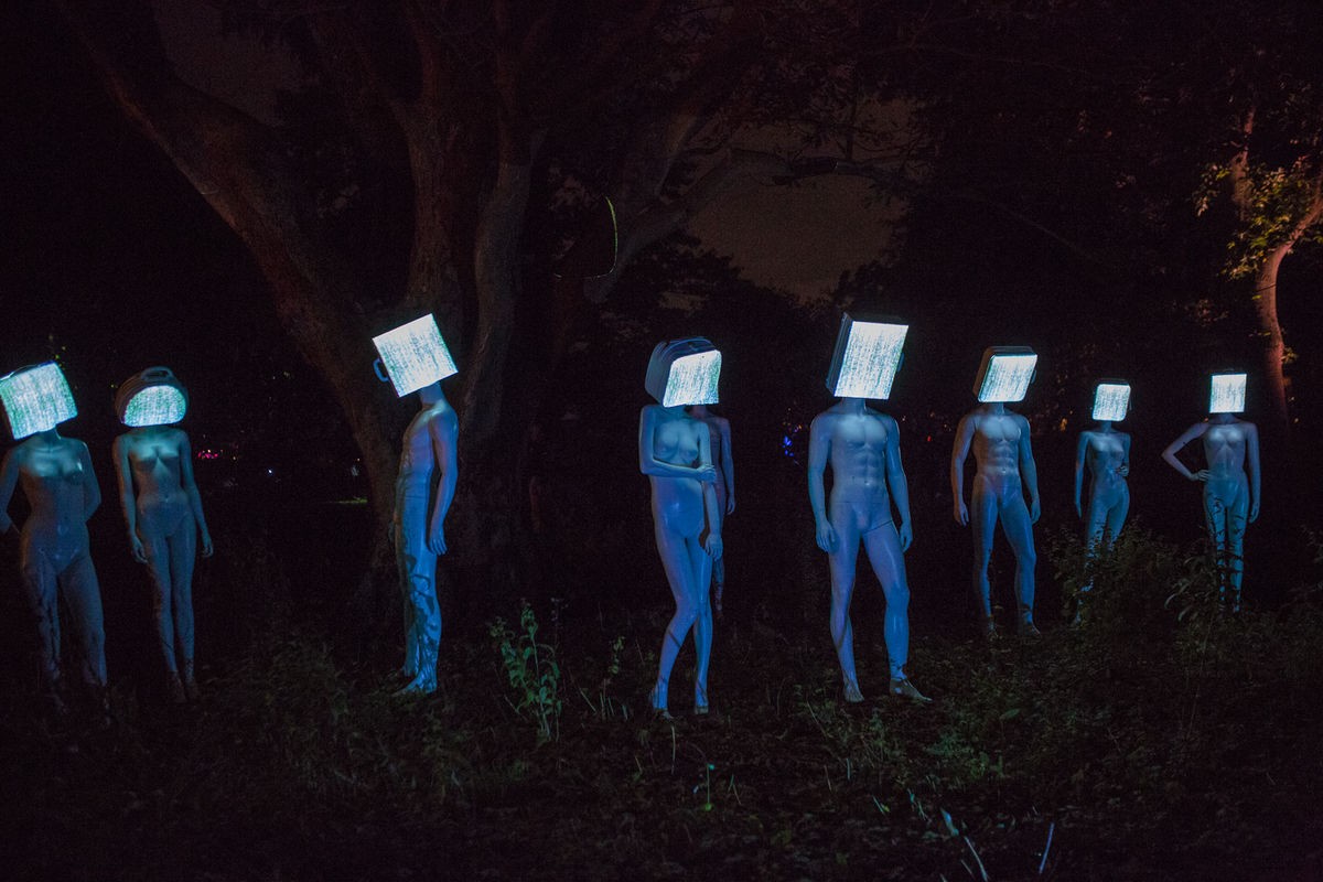 Een kunstwerk. 8 mannequins zijn tegen een donkere achtegrond geposeerd. Hun hoofden zijn bedekt met vierkante lichtgevende blokken.