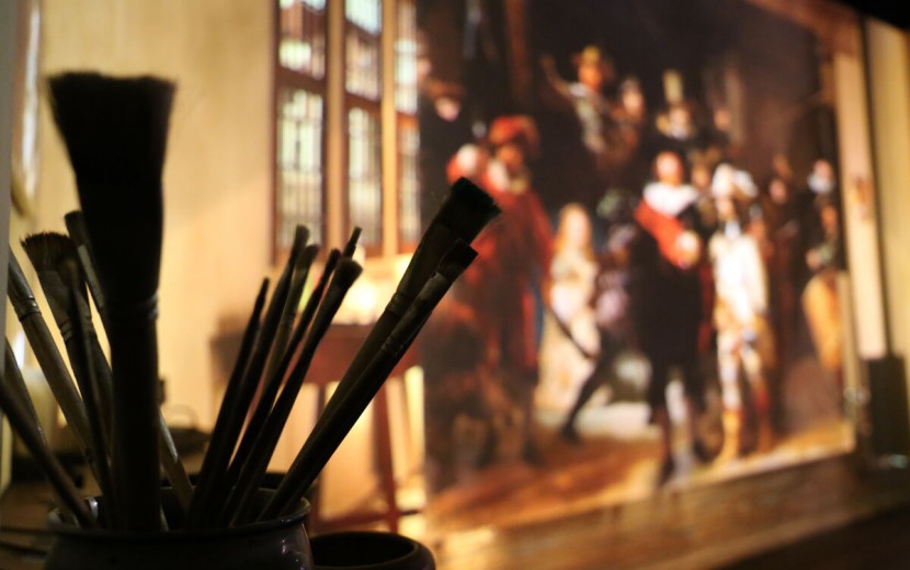 Een close-up van een bundel verfkwasten. In de achtergrond, niet in focus, is een stuk van De Nachtwacht van Rembrandt van Rijn te zien. 
