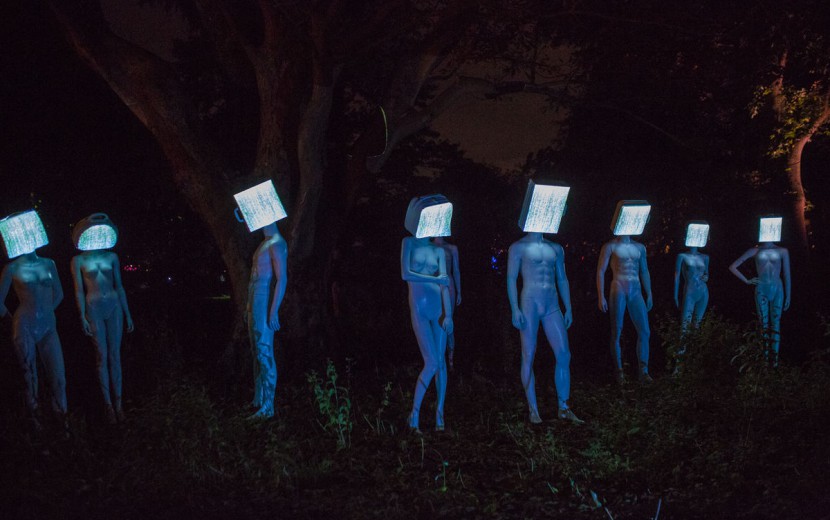Een kunstwerk. 8 mannequins zijn tegen een donkere achtegrond geposeerd. Hun hoofden zijn bedekt met vierkante lichtgevende blokken. 